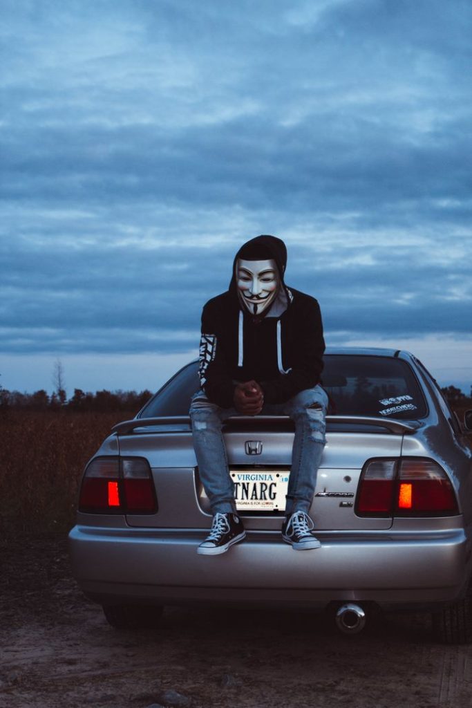 Person wearing an anonymous mask sits on a car trunk during twilight in Williamsburg, VA.