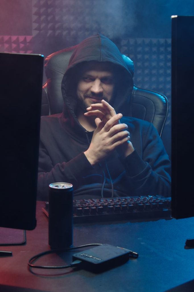 A man in a hoodie sits at a dimly lit desk with a keyboard and computer, evoking a mysterious atmosphere.
